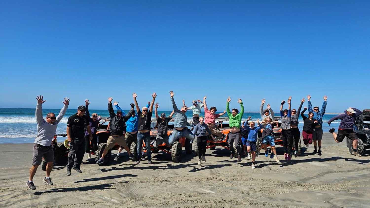 A large tour group leaping in the air on the beach in front of a line of Speed UTVs