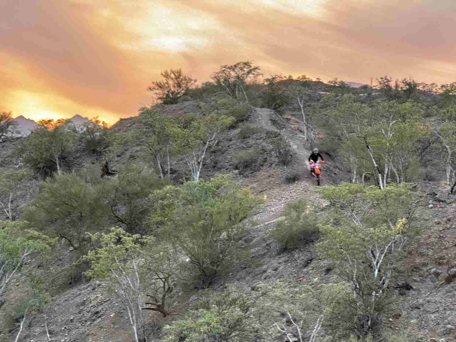 A dirt bike rider on a narrow desert ridge trail under an orange sunset sky in Loreto