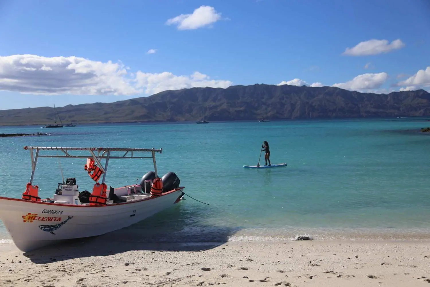 A panga pulled up on a white sand beach while a paddleboarder crosses the turquoise bay